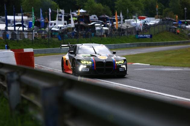 Ein mattschwarzer BMW M4 GT3 Luca Engstler & Mirko Bortolotti & Patric Niederhauser (Red Bull Team ABT) durchfährt eine Kurve auf dem Nürburgring. Das Fahrzeug trägt die charakteristische Red-Bull-Livery mit dem markanten Stier-Logo auf der Flanke sowie den BMW-Motorsport-Farben Blau-Violett-Rot als vertikale Streifen auf der Frontpartie. Sponsoren wie Bosch, H&R und Shell sind auf der Karosserie sichtbar. Im Hintergrund sind Fahnen, Teamzelte und das Fahrerlager des 24h-Rennens zu erkennen. BMW M4 GT3 auf der Strecke am Nürburgring Copyright: xMichaelxGelhardtx
