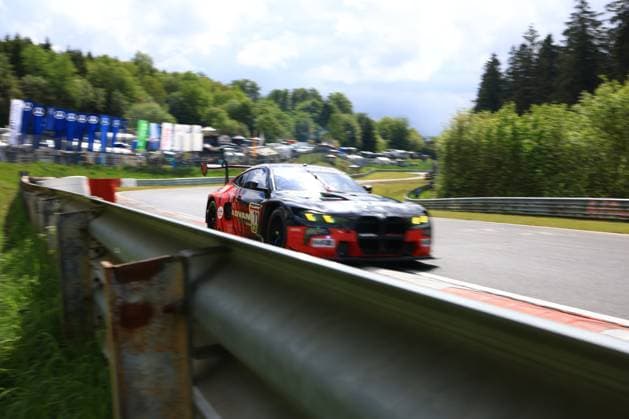Das Giuliano Alesi & Kazuto Kotaka & Miki Koyama & Shunji Okumoto (Toyo Tires with Ring Racing) von Toyo Tires with Ring Racing durchfährt mit hohem Tempo eine Kurve auf der Nordschleife am Nürburgring. Der BMW M4 GT3 zeigt eine auffällige schwarz-rote Livery mit ADVAN-Sponsoring. Im Hintergrund sind die charakteristischen grünen Hügel der Eifel sowie das Fahrerlager zu erkennen. Das Fahrzeug wird durch eine Leitplanke im Vordergrund gerahmt, was die gefährliche Nähe zur Streckenbegrenzung unterstreicht. BMW M4 GT3 170 von Toyo Tires with Ring Racing auf der Nordschleife Copyright: xMichaelxGelhardtx