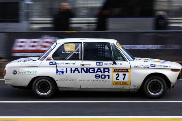 Ein historischer BMW 2002 mit der Startnummer 27 passiert in der Boxengasse des Nürburgrings die Boxenmauer. Das weiße Fahrzeug trägt die Aufschrift Hangar 901 Aircraft Maintenance als Hauptsponsor sowie die Logos von s2-software, RAVENOL und UA-Racing. Im Cockpit ist der Name Bernhard Hagen auf dem Türrahmen zu lesen. Das Fahrzeug nimmt am DHLM (Deutsche Historische Langstrecken-Meisterschaft) teil und zeigt deutliche Gebrauchsspuren vom Renneinsatz. Klassischer BMW 27 mit Hangar 901-Sponsoring in der Boxengasse