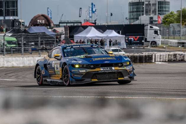 Der Ford Mustang GT3 mit der Startnummer 64 von HRT Ford Racing durchfährt eine Kurve auf dem Nürburgring. Das in Dunkelblau und Gold gehaltene Fahrzeug trägt die Sponsoren RAVENOL, H&R sowie Yokohama und ist mit dem Gran Turismo-Logo auf der Motorhaube versehen. Im Hintergrund sind die charakteristischen Gebäude des Nürburgring-Fahrerlagers sowie Hospitality-Zelte zu erkennen. Christopher Mies & Dennis Olsen & Emil Christian Gjerdrum & Finn Wiebelhaus & Frederic Vervisch (HRT Ford Racing) Ford Mustang GT3 64 von HRT Ford Racing am Nürburgring Copyright: xMichaelxSchäferx