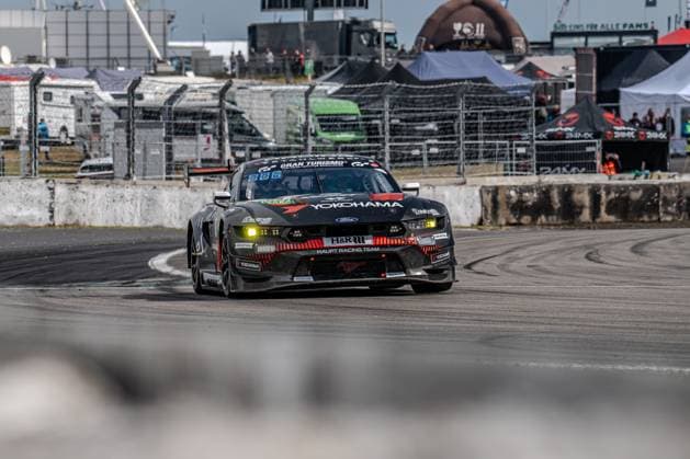Der Ford Mustang GT3 mit der Startnummer 65 des HRT Ford Racing Teams durchfährt eine Kurve am Nürburgring. Das schwarz lackierte Fahrzeug trägt die markanten Yokohama- und H&R-Sponsoren sowie den Gran Turismo-Schriftzug auf dem Dach. Die gelben Bremslichter leuchten beim Einlenken, im Hintergrund sind Absperrungen, Zuschauerbereich und Teamzelte zu erkennen. Colin Caresani & David Schumacher & Hubert Haupt & Vincent Kolb (HRT Ford Racing) Ford Mustang GT3 65 von HRT Ford Racing am Nürburgring Copyright: xMichaelxSchäferx