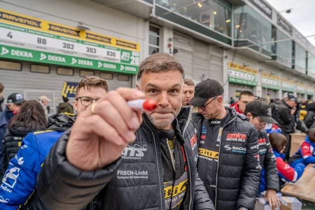 Im Fahrerlager des Nürburgrings hält ein Mann in einer Jacke mit dem Aufdruck FRISCH-Licht Technische LED-Beleuchtung und Giti Competition Tires einen roten Stift direkt in die Kamera. Im Hintergrund sind weitere Personen in Teamkleidung mit dem emka Motor Oils - und SACHS Performance -Logo zu sehen. An den Boxengaragentoren sind RAVENOL- und DEKRA-Banner sowie Startnummern-Schilder (88, 302, 303, 830) erkennbar. Teamvertreter mit Stift im Fahrerlager am Nürburgring Copyright: xMichaelxSchäferx