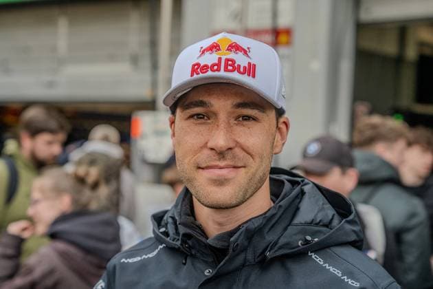 Im Fahrerlager des Nürburgrings ist Kelvin van der Linde zu sehen, der eine Red-Bull-Cap und eine MCG-Teamjacke trägt. Im Hintergrund sind weitere Personen und Teamstrukturen erkennbar, die auf die lebhafte Atmosphäre des 24h-Rennwochenendes hinweisen. Kelvin van der Linde im Fahrerlager am Nürburgring Copyright: xMichaelxSchäferx