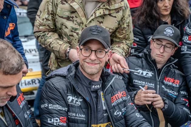 Im Fahrerlager des Nürburgrings versammeln sich mehrere Personen in der Teamkleidung von WS Racing Motorsport. Im Vordergrund ist Jan Ullrich zu sehen, dessen Jacke Sponsoren wie emka Motor Oils, FRISCH-Licht, up2race und WS Racing eSports zeigt. Im Hintergrund sind weitere Teammitglieder sowie Zuschauer zu erkennen. Fahrerlager-Atmosphäre beim 24h Nürburgring mit WS Racing-Team Copyright: xMichaelxSchäferx