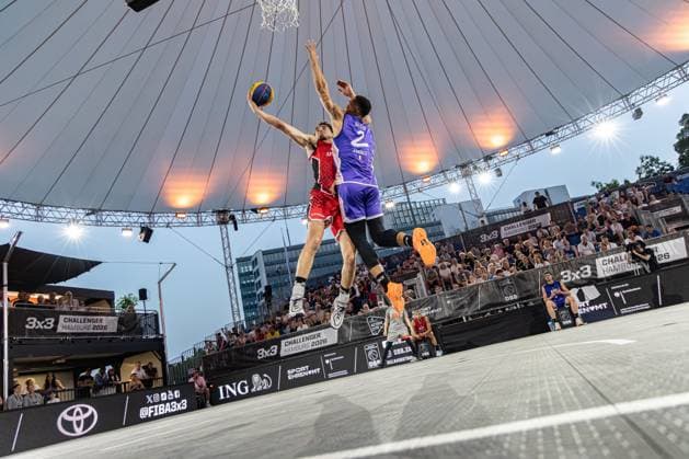 Beim FIBA 3x3 Hamburg Challenger 2026 in der Active City Arena zeigt sich eine intensive Spielsituation: Ein Spieler im roten Trikot versucht einen Dunk, während sein Gegenspieler ( 2, Toulouse FRA) im blauen Trikot den Wurf blockt. Das Spiel findet unter freiem Himmel mit beeindruckender Zeltdach-Arena statt. Spektakulärer Dunk-Versuch gegen Toulouse in der Active City Arena Hamburg