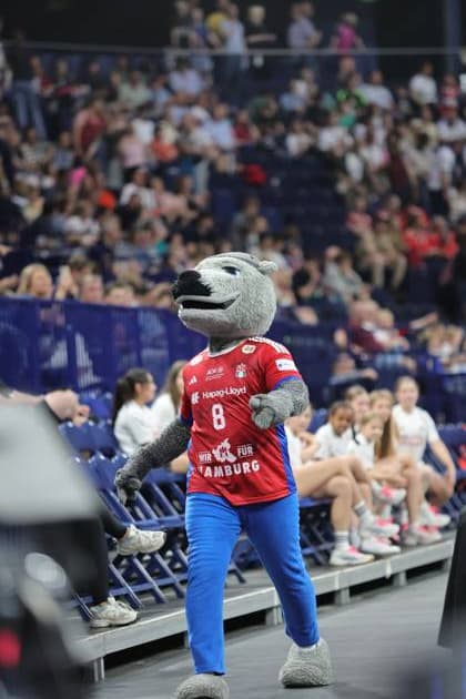 Das HSV-Maskottchen in rotem und blauem Kostüm winkt den Zuschauern in der Barclays Arena zu. Im Hintergrund sind Spieler in blauen HSV-Trikots und Fans in den Rängen zu sehen. HSV Hamburg Maskottchen bei Spielerbetreuung