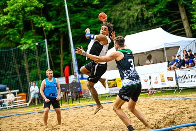 Ein Spieler im weißen Trikot springt hoch und wirft den Ball, während Spieler 3 im schwarz-grünen Trikot mit dem Aufdruck Rotterdam Topsport versucht, den Wurf zu blocken. Im Hintergrund beobachtet ein weiterer Spieler im blauen Trikot die Szene auf dem Sandplatz. Spektakulärer Sprungwurf beim Beach-Handball-Turnier