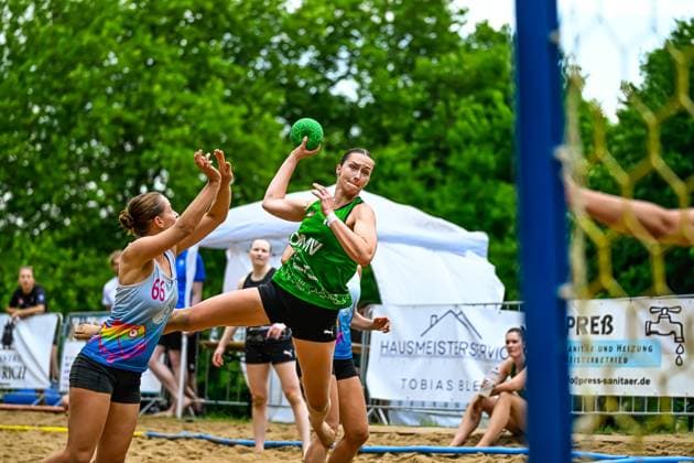 Eine Spielerin im grünen Trikot zieht aus dem Sprung zum Wurf ab, während Spielerin 66 im bunten Trikot die Abwehr versucht. Das Spiel findet auf einem Sandplatz im Freien statt. Wurfaktion beim Beach-Handball: Spielerin im grünen Trikot zieht ab