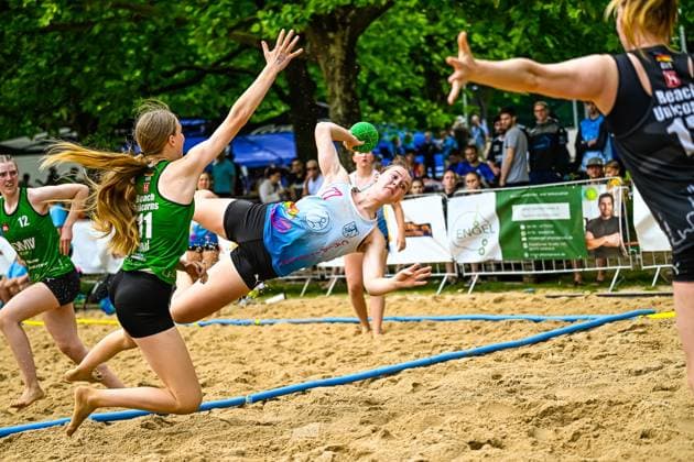 Eine Spielerin im hellblauen Trikot wirft sich in einer akrobatischen Drehbewegung mit dem Ball in der Hand über die Spielfeldlinie. Zwei Spielerinnen der Beach Unicorns ( 11 und 12) versuchen den Wurf zu blocken, während 15 der Beach Unicorns als Torfrau bereitsteht. Spektakulärer Wurfversuch im Beach-Handball-Spiel gegen Beach Unicorns