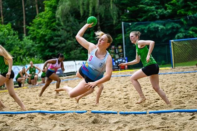 Eine Spielerin mit der Nummer 15 im weißen Trikot springt beim Beach-Handball-Spiel in die Luft und wirft den grünen Ball aufs Tor. Gegenspielerinnen in grünen Trikots versuchen den Angriff zu unterbinden. Spielerin ( 15) beim Wurfsprung im Beach-Handball