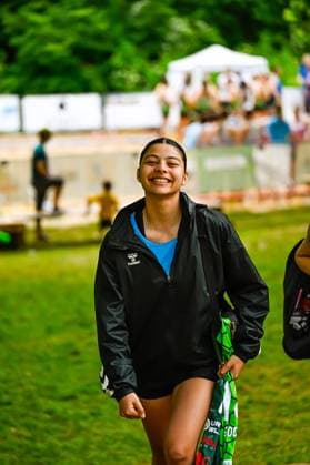 Eine junge Spielerin in schwarzer Hummel-Trainingsjacke und blauem Unterziehshirt läuft lächelnd über eine Rasenfläche. Sie trägt ein grünes Torwarttrikot in der Hand. Lächelnde Spielerin mit Hummel-Jacke bei Outdoor-Sportveranstaltung