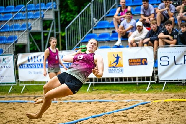 Eine Spielerin mit der Nummer 15 im pinken Trikot wirft beim Karacho Beach Cup German Beach Open in der Luft schwebend auf das Tor. Im Hintergrund verfolgen Zuschauer auf blauen Tribünensitzen das Spiel. Spielerin ( 15) beim Wurfversuch beim Karacho Beach Cup