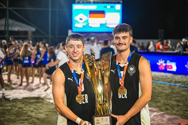 Zwei Spieler der deutschen Beachhandball-Nationalmannschaft halten gemeinsam den goldenen Siegerpokal und tragen Goldmedaillen um den Hals. Im Hintergrund ist die Anzeigetafel mit der deutschen Flagge sowie feiernde Zuschauer am Jarun See in Zagreb zu sehen. IHF Beach Handball World Championship, WM, Weltmeisterschaft 2026, 23.06.2026, Zagreb, Jarun See