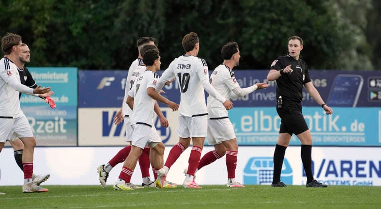 Foto : JUbel bei Drochtersen nach dem verrückten Torschuss ( Bogenlampe ) von Christopher SCHEPP , hier protestieren die Phönix Spieler Fabio Oliveira MAIOLO und Jonathan STÖVER ( 10 ) und Benjamin LUIS ( 24 ) gegen Schiri Dominik Kopmann Fussball Regionalliga Nord, 1. Spieltag am Fr. 31.07.2026 SV Drochtersen - Phönix Lübeck 2 - 1