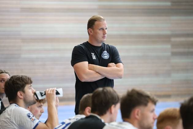 VfL Lübeck-Schwartau-Trainer David Röhrig steht mit verschränkten Armen konzentriert an der Seitenlinie und verfolgt das Spiel in der Hansehalle Lübeck. Im Hintergrund sitzen mehrere Spieler auf der Auswechselbank, darunter Guy Resch Ermann. David Röhrig beobachtet das Spielgeschehen von der Seitenlinie