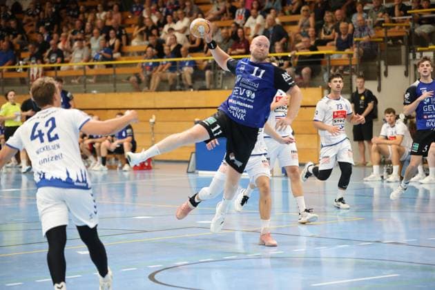 Kristoffer Jessen ( 11; Grindstedt GIF) zieht aus dem Sprung ab und hält den Ball wurfbereit über dem Kopf, während Lukas Kingsley Emdorf ( 73; VfL Lübeck-Schwartau) und Einar Nickelsen ( 42; VfL Lübeck-Schwartau) die Szene beobachten. Im Hintergrund verfolgen weitere Spieler beider Teams sowie eine gut gefüllte Tribüne das Geschehen. Kristoffer Jessen ( 11; Grindstedt GIF) beim Sprungwurf in der Hansehalle