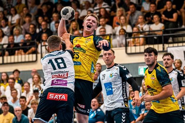 Haukur Thrastarson ( 25; Rhein Neckar Löwen) springt im Zweikampf mit Felix Kazmeier ( 18; SV Salamander Kornwestheim) zum Wurf hoch und hält den Ball über dem Kopf. Im Hintergrund beobachten Jan Döll ( 29; SV Salamander Kornwestheim) und weitere Spieler beider Teams die Szene in der vollbesetzten Sporthalle Ost Kornwestheim. Haukur Thrastarson ( 25; Rhein Neckar Löwen) zieht zum Wurf hoch