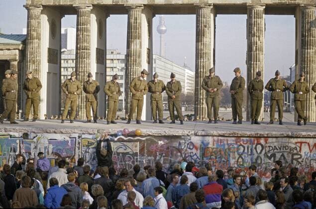 Grenzer auf der Berliner Mauer und Menschenmassen davor am Brandenburger Tor während des Mauerfalls iblnmi00182061