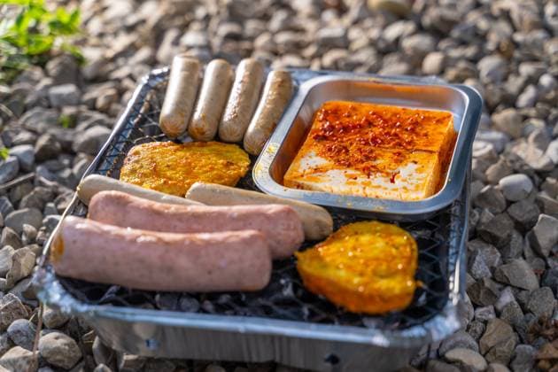 Vegetarisch Grillen, Bratwürste Vegan und Vegetarisch auf einem Einweggrill auf Kieselsteinen neben einer Wiese. Fleisch und Wurst Ersatzprodukte