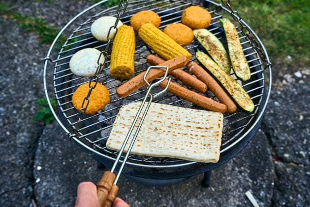 Ein bunter Mix aus vegetarischen Köstlichkeiten wie Maiskolben, vegane Würstchen, Zucchini und Grillkäse brutzeln auf einem Kohlegrill. Ein Mann wendet mit einer Zange eine fleischlose Bratwurst