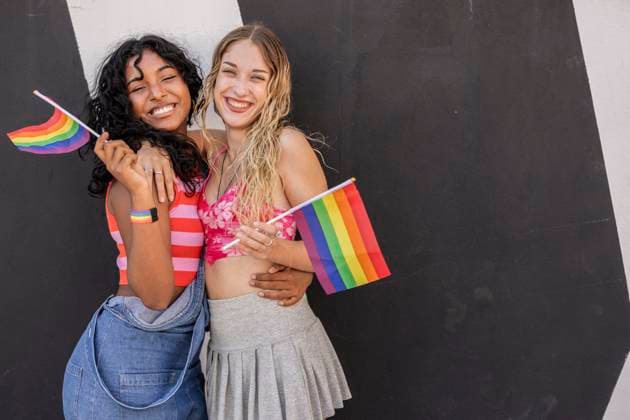 A joyful multiracial lesbian couple embraces while holding LGBTQIA+ pride flags against a contrasting black and white background, symbolizing love and diversity Model Released JoseCarlosIchiro_ID23778_690111_007 Copyright: xJosexCarlosxIchirox