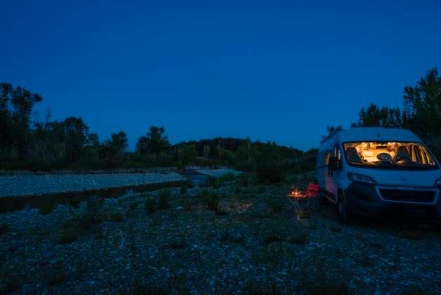 EIn Mann sitzt auf einem Campingstuhl vor einem Tisch an einem Sommerabend zur blauen Stunde vor seinem Camper. Deutschland Mit einem Campervan unterwegs