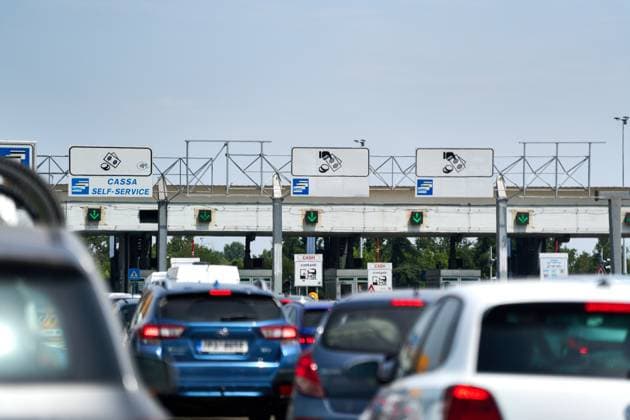 Fahrzeuge warten in einer Schlange und großem Andrang vor einer Mautschranke mit klar sichtbaren Symbolen für Bargeld- und Kartenzahlung. Grüne Signale zeigen geöffnete Spuren.