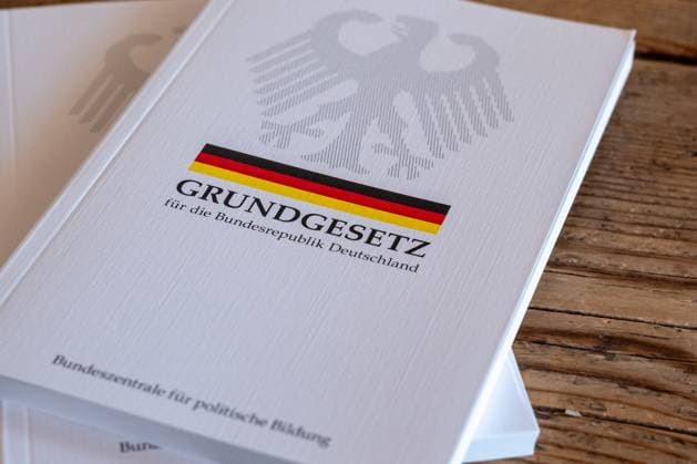 Grundgesetz der Bundesrepublik Deutschland auf Holztisch 19.06.25, Limburg: Symbolfoto, Illustrationsbild, Symbolbild, Illustrationsfoto, Alltagsszene Ein gedrucktes Exemplar des Grundgesetzes der Bundesrepublik Deutschland liegt auf einem rustikalen Holztisch. Die Ausgabe mit Bundesadler, schwarz-rot-goldenem Streifen und dem Titel Grundgesetz für die Bundesrepublik Deutschland symbolisiert demokratische Werte, Rechtsstaatlichkeit und politische Bildung in Deutschland. Im Hintergrund ist ein zweites Exemplar sichtbar. Limburg Copyright: xW2Artx xKlaraxMichelx