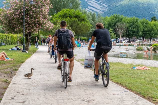 Am Uferweg des Gardasees bei Torbole fahren Urlauber mit dem Fahrrad an Badestellen vorbei. Der beliebte Ort ist im Sommer gut besucht, Radwege teilen sich Touristen mit Spaziergängern und Tieren