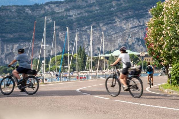 Radfahrerinnen und Radfahrer auf der Promenade am Gardasee unterwegs mit Helm und Gepäck entlang der Uferstraße, wo See und Berge eine aktive Urlaubsstrecke bieten.