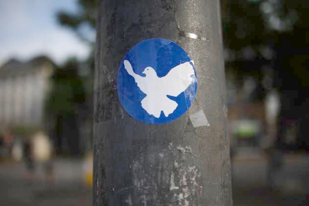 Flensburg, Schleswig-Holstein Auf einem grauen, leicht verwitterten Laternenpfahl ist ein runder blauer Aufkleber mit einer weißen Friedenstaube zu sehen. Die Taube ist fliegend dargestellt und symbolisiert Frieden. Der Hintergrund des Bildes ist unscharf, was auf eine belebte städtische Umgebung hinweist. Die Aufnahme wirkt dokumentarisch und vermittelt eine friedliche Botschaft in einem urbanen Kontext. Aufnahme vom 20.07.2025, Flensburg, Innenstadt