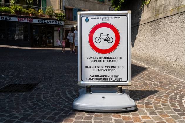 Mobiles Verkehrsschild in Limone sul Garda: roter Kreis mit Fahrrad, Hinweis: Fahrräder nur mit Handführung erlaubt. Auf dem kopfsteinigen Altstadt-Platz passieren Fußgänger. Regel für die Fußgängerzone