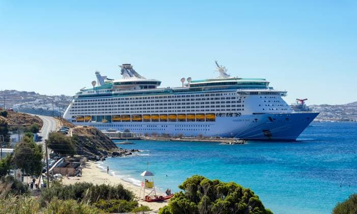 Mykonos, Kykladen, Griechenland - Kreuzfahrtschiff Voyager of the Seas liegt in der Hafenbucht Mykonos. Vorne baden Touristen am Sandstrand Paralia Agios Stefanos. Hinten die Altstadt von Mykonos. Mykonos Mykonos Griechenland