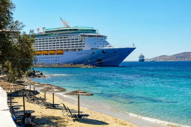 Mykonos, Kykladen, Griechenland - Kreuzfahrtschiff Voyager of the Seas liegt in der Hafenbucht Mykonos. Vorne Strandliegen und Sonnenschirme am Sandstrand Paralia Agios Stefanos. Mykonos Mykonos Griechenland