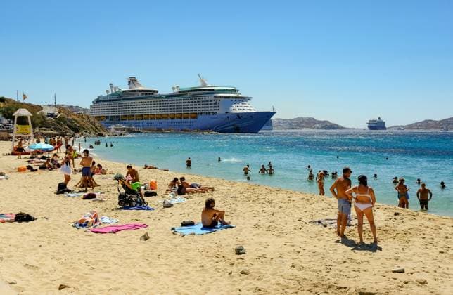 Mykonos, Kykladen, Griechenland - Kreuzfahrtschiff Voyager of the Seas liegt in der Hafenbucht Mykonos. Vorne baden Touristen und Einheimische am Sandstrand Paralia Agios Stefanos. Mykonos Mykonos Griechenland