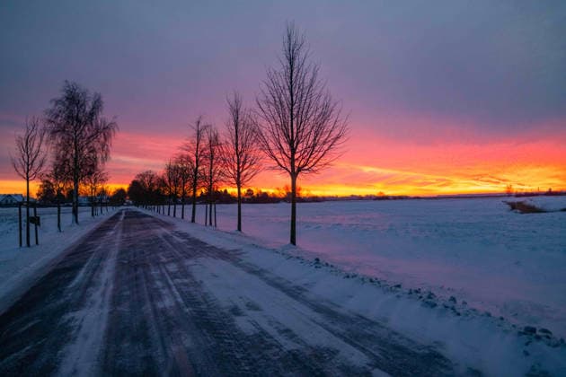 Rostock, Deutschland 09. Januar 2026: Im Bild: Eine schmale Landstraße verläuft durch eine winterliche Landschaft im Landkreis Rostock. Der Fahrbahnrand und die angrenzenden Felder sind mit Schnee bedeckt, entlang der Straße stehen kahle Bäume. Am Horizont färbt ein intensiver Sonnenauf- oder Sonnenuntergang den Himmel in Orange-, Rot- und Violetttönen und sorgt für eine markante Lichtstimmung in der kalten Jahreszeit. Mecklenburg Vorpommern Copyright: xFotostandx xVoelkerx