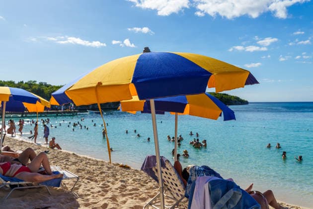 Unter bunten Sonnenschirmen liegen Urlauber im Schatten, während andere im klaren Meer baden und sich sonnen. Die Szene zeigt den typischen Sommerurlaub am Strand mit Erholung, Abkühlung und sonniger Ferienstimmung.