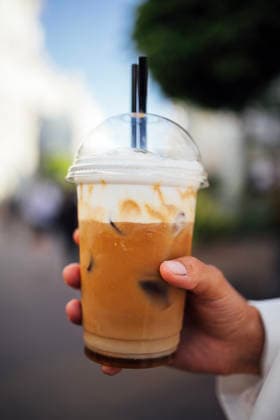 IMAGO Creative, Food, Trend, Iced Coffee Eiskaffee in einem durchsichtigen Plastikbecher, gehalten von einer Hand mit zwei schwarzen Strohhalmen, verschwommener städtischer Hintergrund mit Grünflächen und vorbeigehenden Menschen Iced coffee drink in a clear cup with whipped cream and two straws, held by a hand against a blurred urban setting with greenery and pedestrians (License=RF) Copyright: xZoonar.com DashaxPetrenkox 24722646