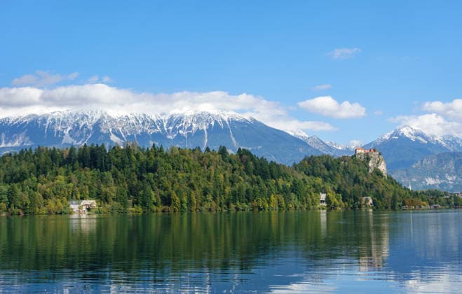 Panoramablick auf den Bleder See mit seiner ikonischen Burg auf einer Klippe und schneebedeckten Alpengipfeln. Ein Symbol für Naturschutz, alpine Biodiversität und nachhaltigen Tourismus in einer unberührten Umgebung