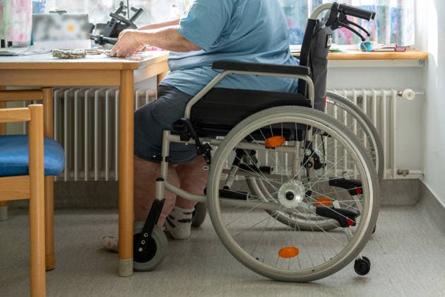 Ein älterer Mensch im Rollstuhl am Tisch verdeutlicht die Herausforderungen im Pflegealltag. Das Bild thematisiert Inklusion, Barrierefreiheit und die notwendige Unterstützung für ein würdevolles Leben.