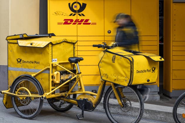 Ein Zustellfahrrad der Deutschen Post steht bereit, während ein Postbote Sendungen zustellt und transportiert. Die Nutzung von Lastenrädern ermöglicht flexible und nachhaltige Zustellung im urbanen Raum.
