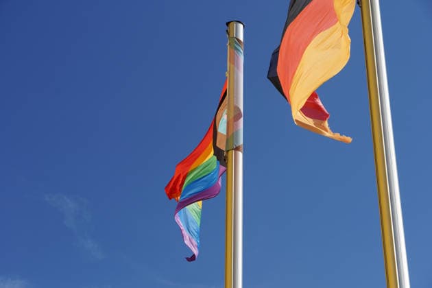 Progress-Pride-Flagge, Progressive Regenbogenfahne und Flagge der Bundesrepublik Deutschland an zwei Fahnenmasten nebeneinander, Symbol für Vielfalt, Toleranz und der Repräsentation von LGBTQIA+-Rechten, queere Menschen als Teil der Gesellschaft