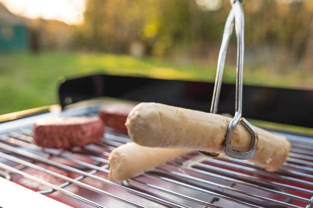 Beim Grillen im Garten im Frühling wendet eine Zange vegane Bratwürste und pflanzliche Steaks auf dem Elektrogrill. Fleischersatzprodukte liegen als Alternative zu klassischem Grillgut auf dem Rost