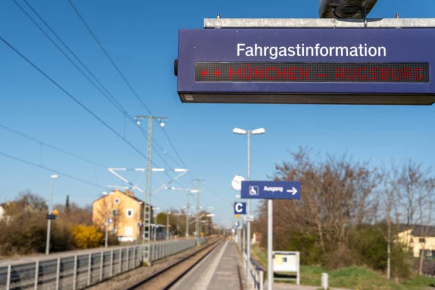 FOTOMONTAGE, Digitale Fahrgastinformation am Bahnhof mit der Anzeige München Augsburg. Das Bild zeigt eine wichtige Pendlerstrecke in Bayern und steht für dichte Zugverbindungen sowie mögliche Störungen im regionalen Bahnverkehr.