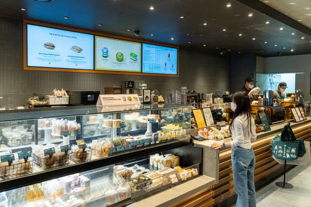 Kundin bestellt an der Theke einer Starbucks-Filiale, während hinter der Glasvitrine Backwaren und Snacks ausgestellt sind und Baristas Getränke zubereiten