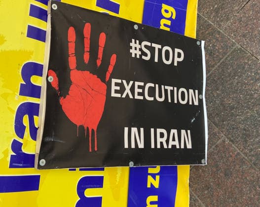 Flensburg, Schleswig-Holstein Zu sehen ist ein schwarzes Protestplakat mit roter Handfläche und der Aufschrift STOP EXECUTION IN IRAN, befestigt vor einem gelben Banner. Die Bildsprache ist klar, direkt und auf visuelle Aufmerksamkeit ausgelegt; sie verweist auf Menschenrechtsverletzungen, Todesstrafe und internationalen Protest. Aufnahme vom 02.05.2026, Flensburg
