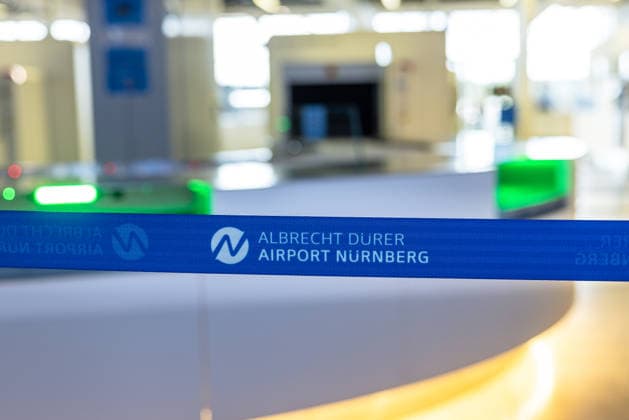 Eröffnung der neuen CT-Kontrollstelle am Airport Nürnberg, Nürnberg, 08.05.2026 Absperrband mit Logo des Albrecht Dürer Airport Nürnberg vor der neuen CT-Kontrollstelle. Im Hintergrund sind unscharf Bereiche der Fluggastkontrolle mit moderner Scannertechnik zu sehen. Nürnberg Bayern Deutschland