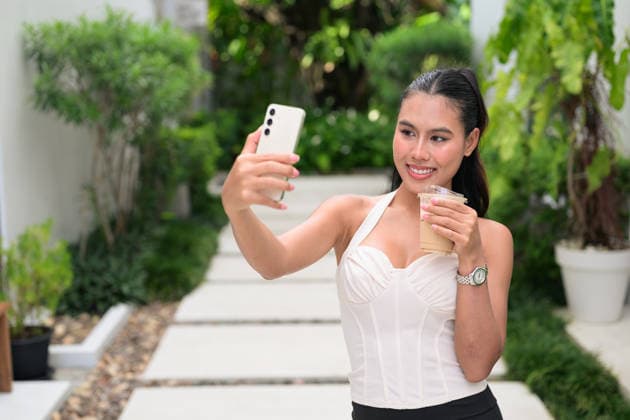 IMAGO Creative, Food, Trend, Iced Coffee Ein fröhlicher Influencer aus dem thailändischen Isan, der während der Sommerferien im Garten mit seinem Handy einen Vlog über Eiskaffee dreht Thai woman from the Isan region vlogging on a white stone walkway. The beautiful Isaan lady wears a cream corset halter blouse while holding a smartphone and iced coffee. That attractive influencer radiates pure happiness while standing outdoors in a luxury resort. Botanical backdrop showcases lush tropical foliage and dense shrubs under bright natural daylight. A stylish brunette fashion model records content for her digital audience. Ivory bodice displays ribbed textures and a structured sweetheart neckline. A fashionable subject stands amidst vibrant greenery in a serene garden environment. This g Copyright: xZoonar.com TonixRantalax 25043796