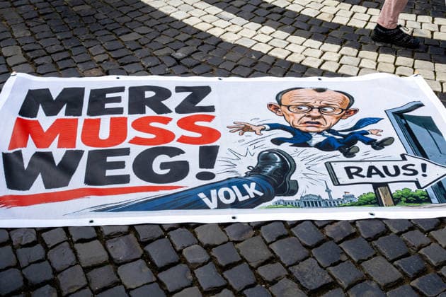 Demonstration gegen die aktuelle Bundesregierung unter Bundeskanzler Friedrich Merz in Augsburg. Unter dem Motto: Merz muss weg! Banner mit Aufschrift: MERZ MUSS WEG! Mit Illustration von einem Fuß auf dem VOLK steht und einer Illustration von Bundeskanzler Friedrich Merz (CDU) der durch eine Tür fliegt mit einem Schild mit Aufschrift: RAUS!