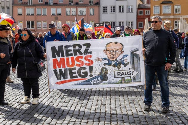 Demonstration gegen die aktuelle Bundesregierung unter Bundeskanzler Friedrich Merz in Augsburg. Unter dem Motto: Merz muss weg! Teilnehmer mit Banner mit Aufschrift: MERZ MUSS WEG! Mit Illustration von einem Fuß auf dem VOLK steht und einer Illustration von Bundeskanzler Friedrich Merz (CDU) der durch eine Tür fliegt mit einem Schild m